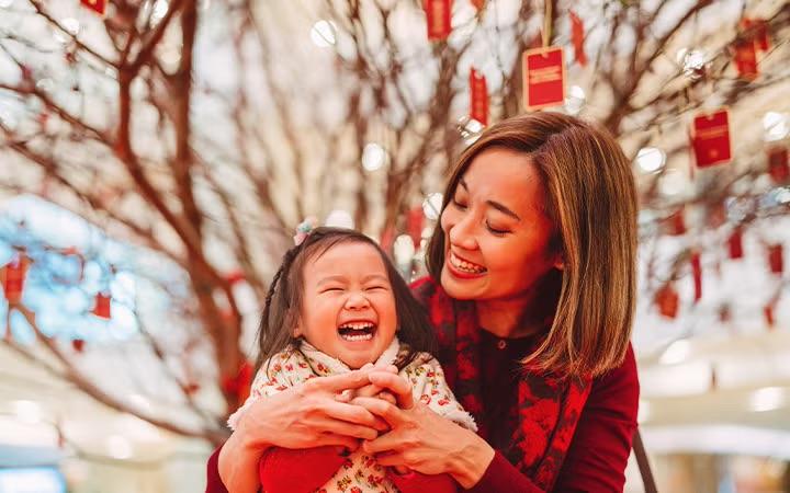 A joyful mother and child celebrate Lunar New Year, surrounded by a tree decorated with red envelopes, symbolising prosperity and business customs.